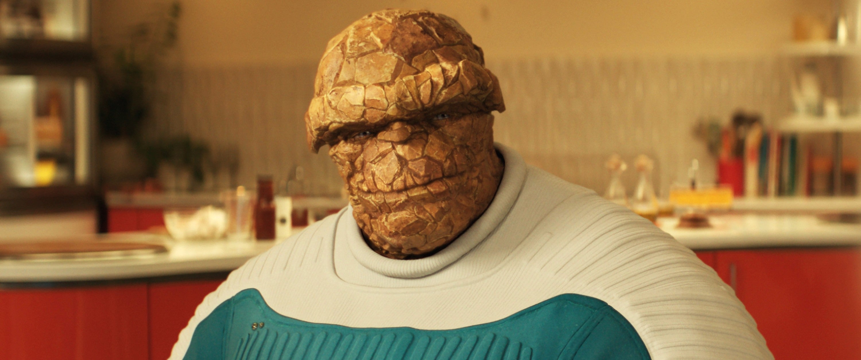 A person in a costume resembling a rock-skinned creature sits in a kitchen setting