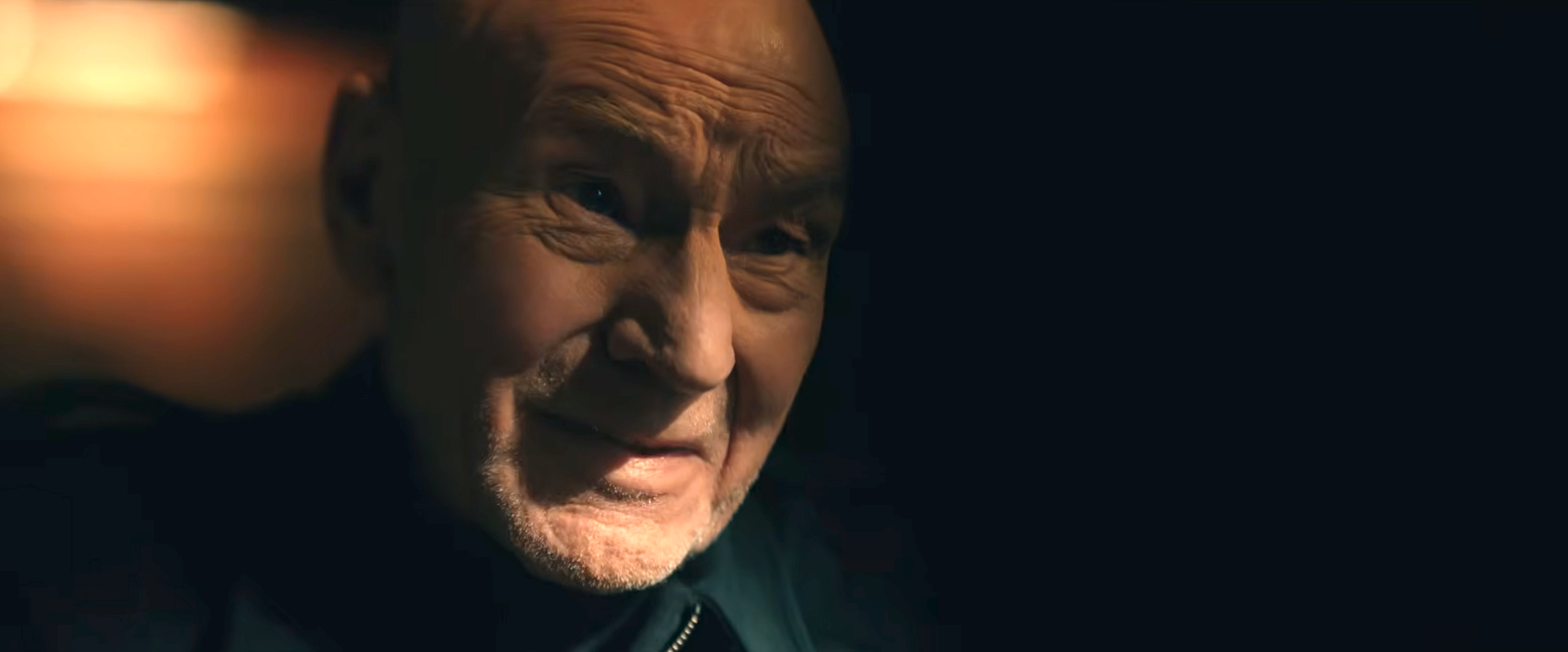 Elderly person in thoughtful expression, face partially shadowed, wearing a dark jacket against a dark background