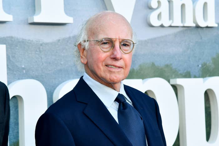 An older person in a classic suit and tie poses at an event, standing in front of a large backdrop with partial text visible