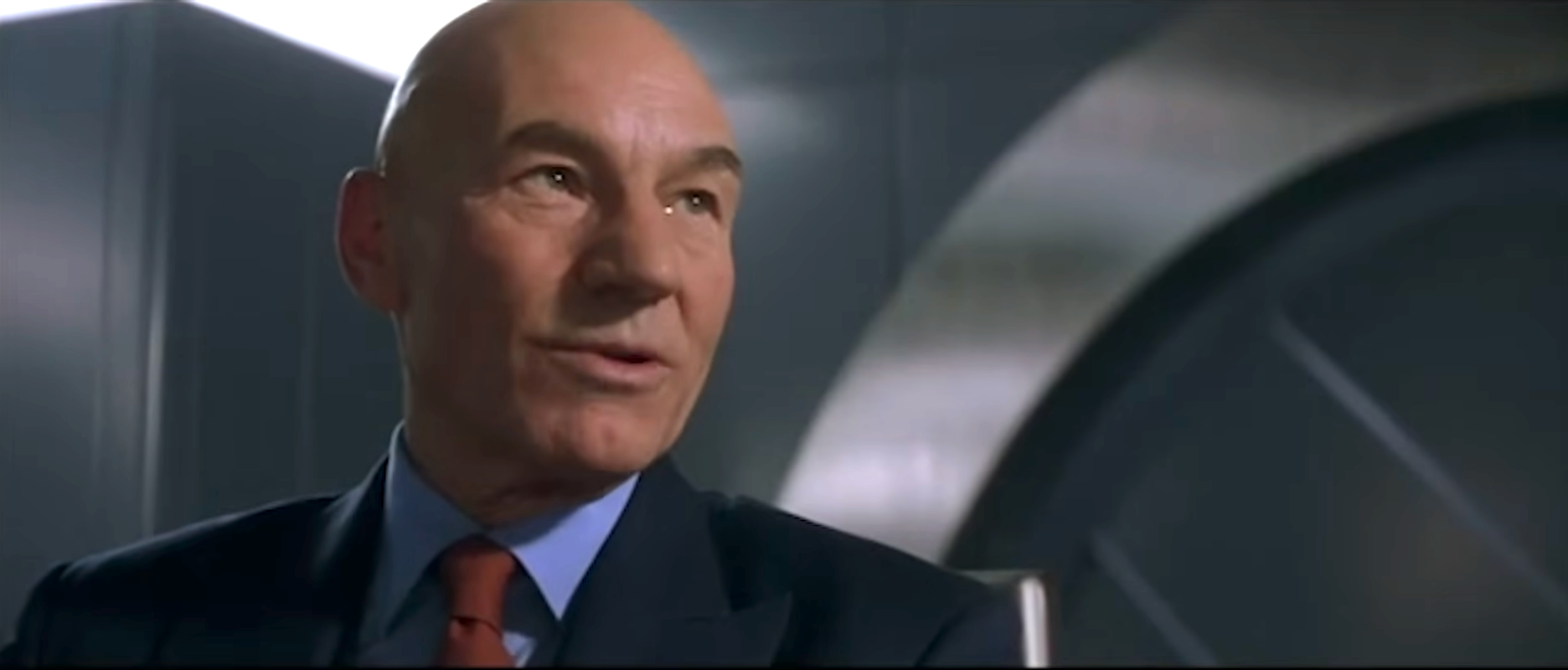 A man in a suit, sitting indoors, speaking