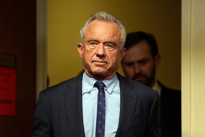 A man in a suit with a blue shirt and patterned tie stands in a doorway, looking serious. Another person is partially visible behind him
