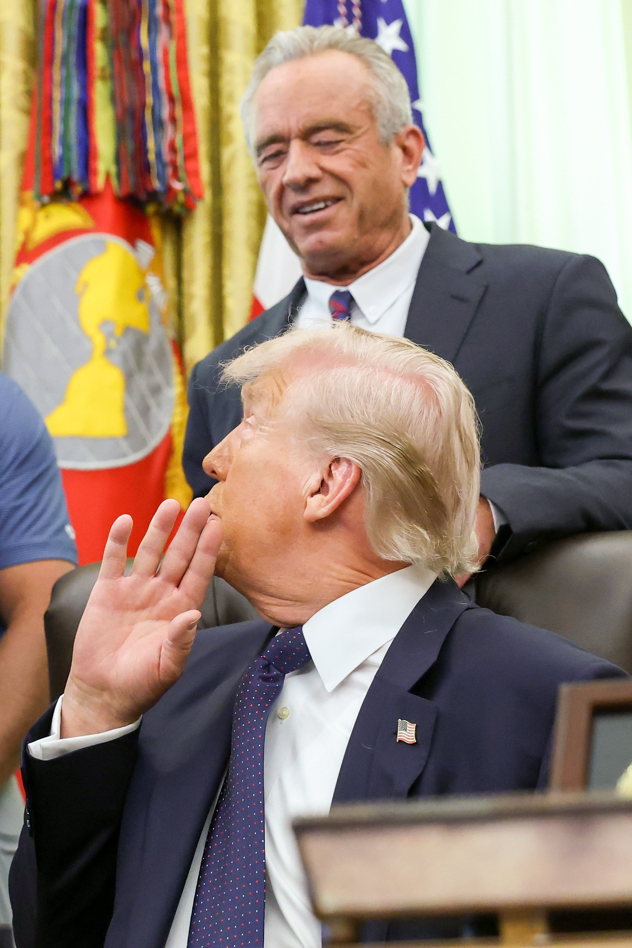 Two men in suits are discussing in an office setting with flags in the background