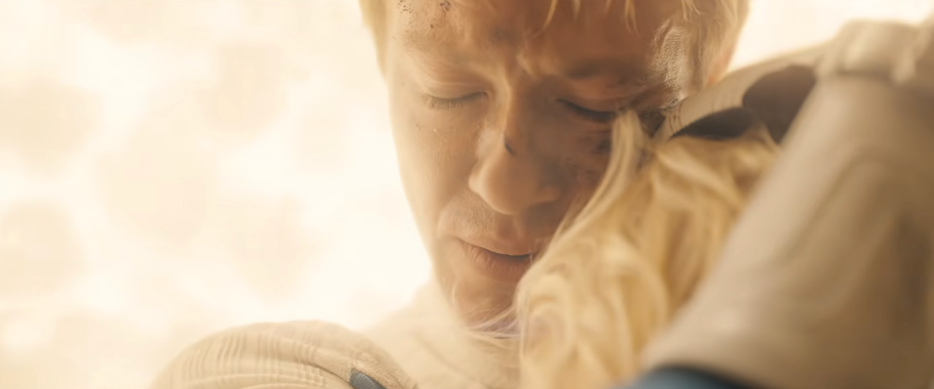 Person in a dramatic scene, eyes closed, embracing another character wearing an armor-like outfit