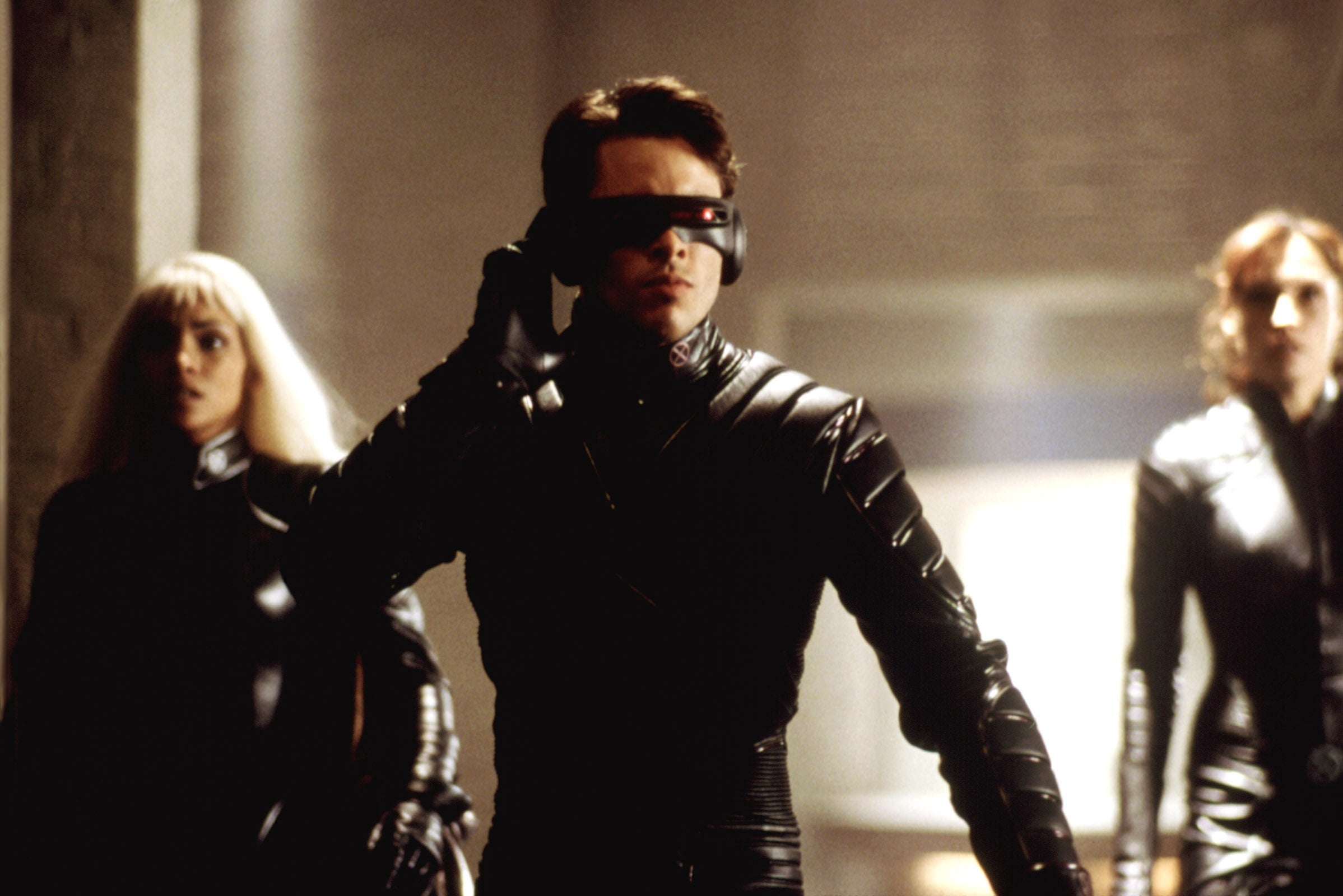 A group in futuristic black leather attire with one person adjusting a visor, resembling a scene from a sci-fi film