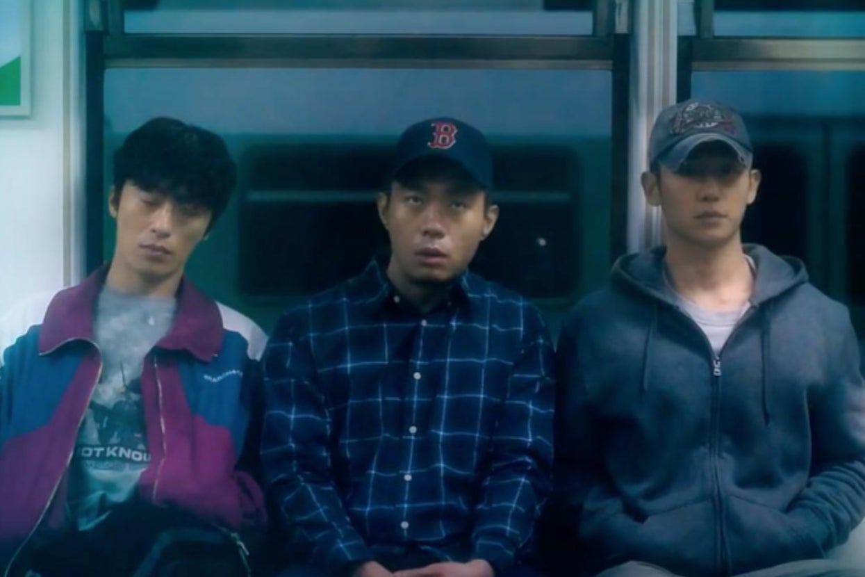 Three men are sitting in a row on a train. The middle one wears a baseball cap and a checkered shirt, flanked by men in casual jackets