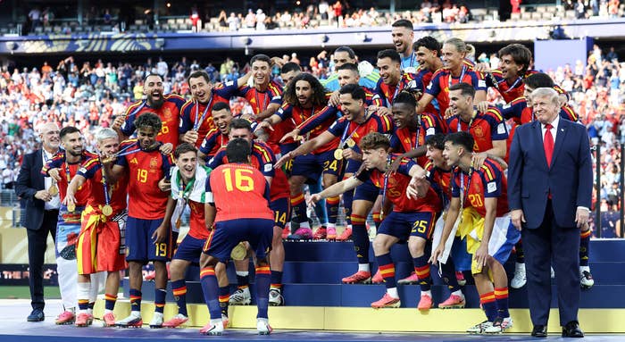 Soccer team celebrating on podium with "FIFA World Cup 2026 Champions" banner. Players wear matching sporty uniforms, posing with enthusiasm