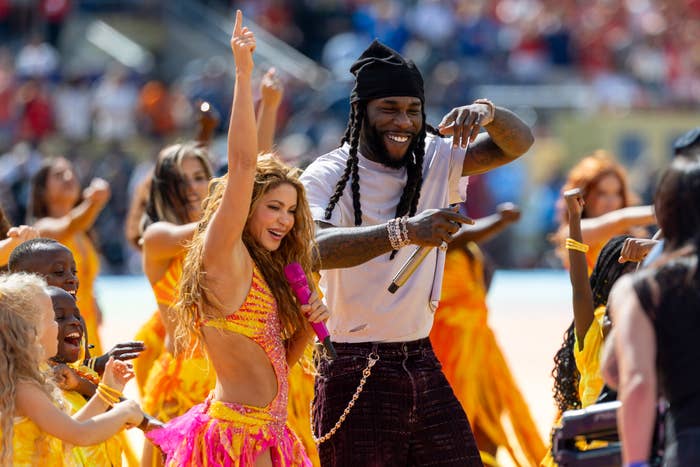 Smiling performers, including a singer in a bright, intricate outfit, dancing energetically on stage with a live audience