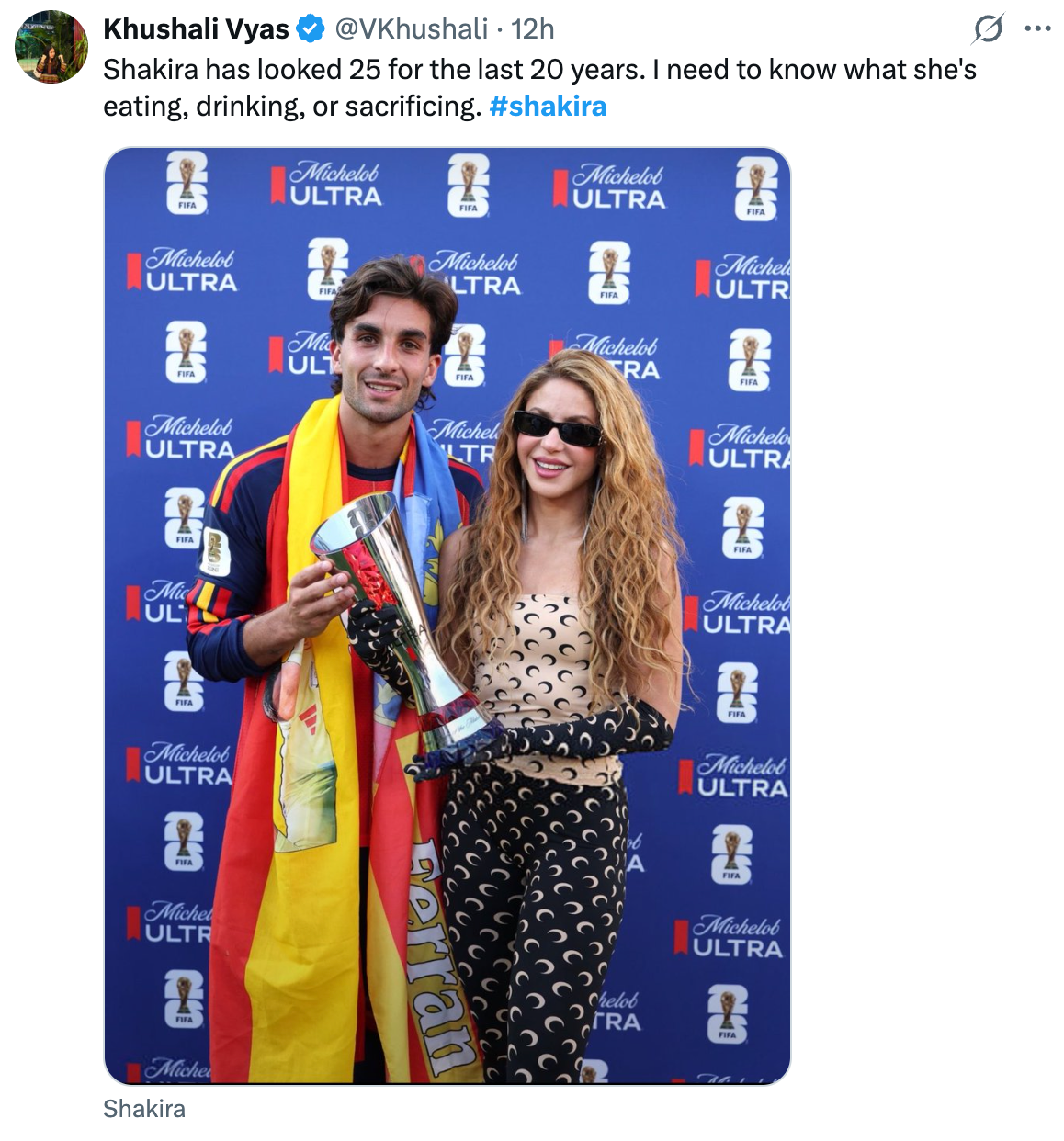 Two people stand together. The woman in a patterned outfit holds a trophy, both smile, with a branded backdrop