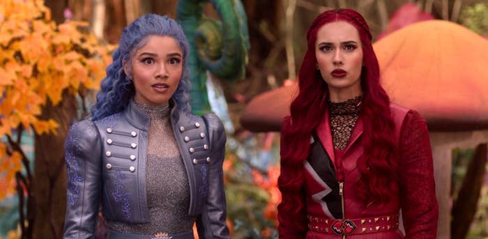 Two women with unique hairstyles and detailed fantasy costumes look surprised in a forest setting