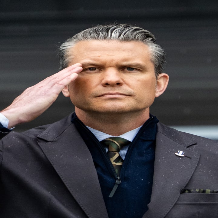 A person in a suit salutes with a serious expression, standing outside against a blurred background