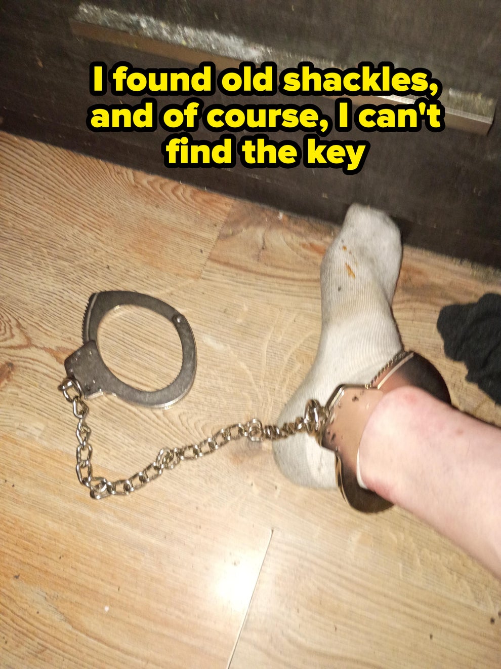 A person's ankle is secured in a handcuff attached to a chain. The floor is wooden, and the person is wearing a sock
