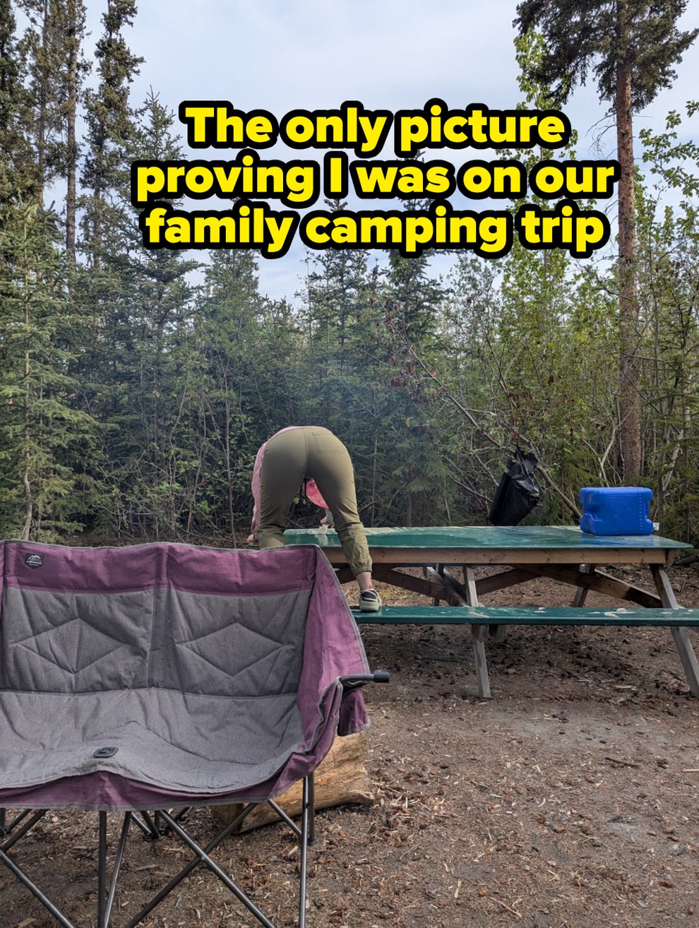 A person is bending over a picnic table in a forested campsite, with a camping chair and gear nearby