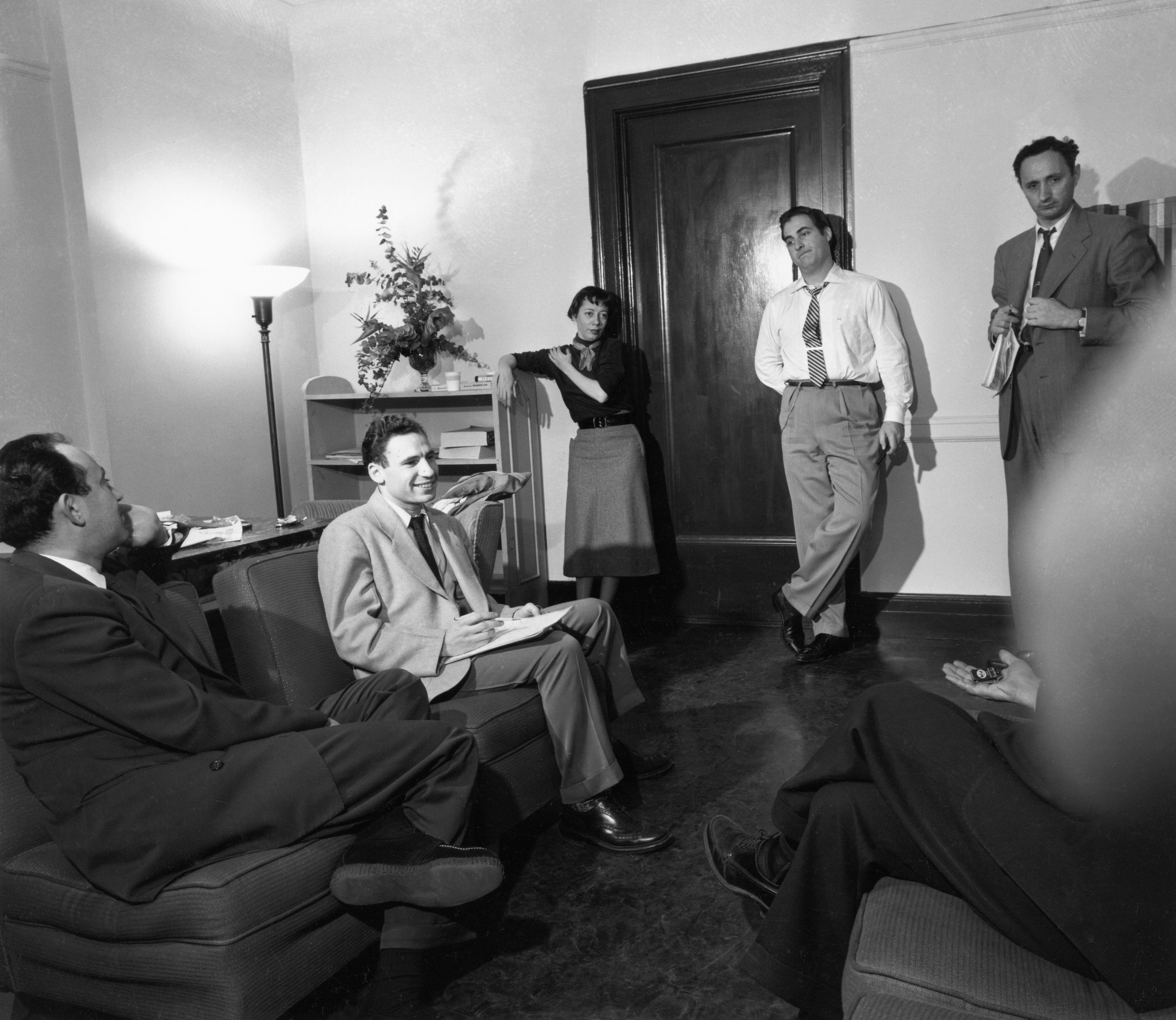 Five people in a vintage office setting, engaged in conversation, with one leaning against a doorframe. Formal mid-20th-century attire. Mel Brooks sits on a couch with a writing pad in his lap