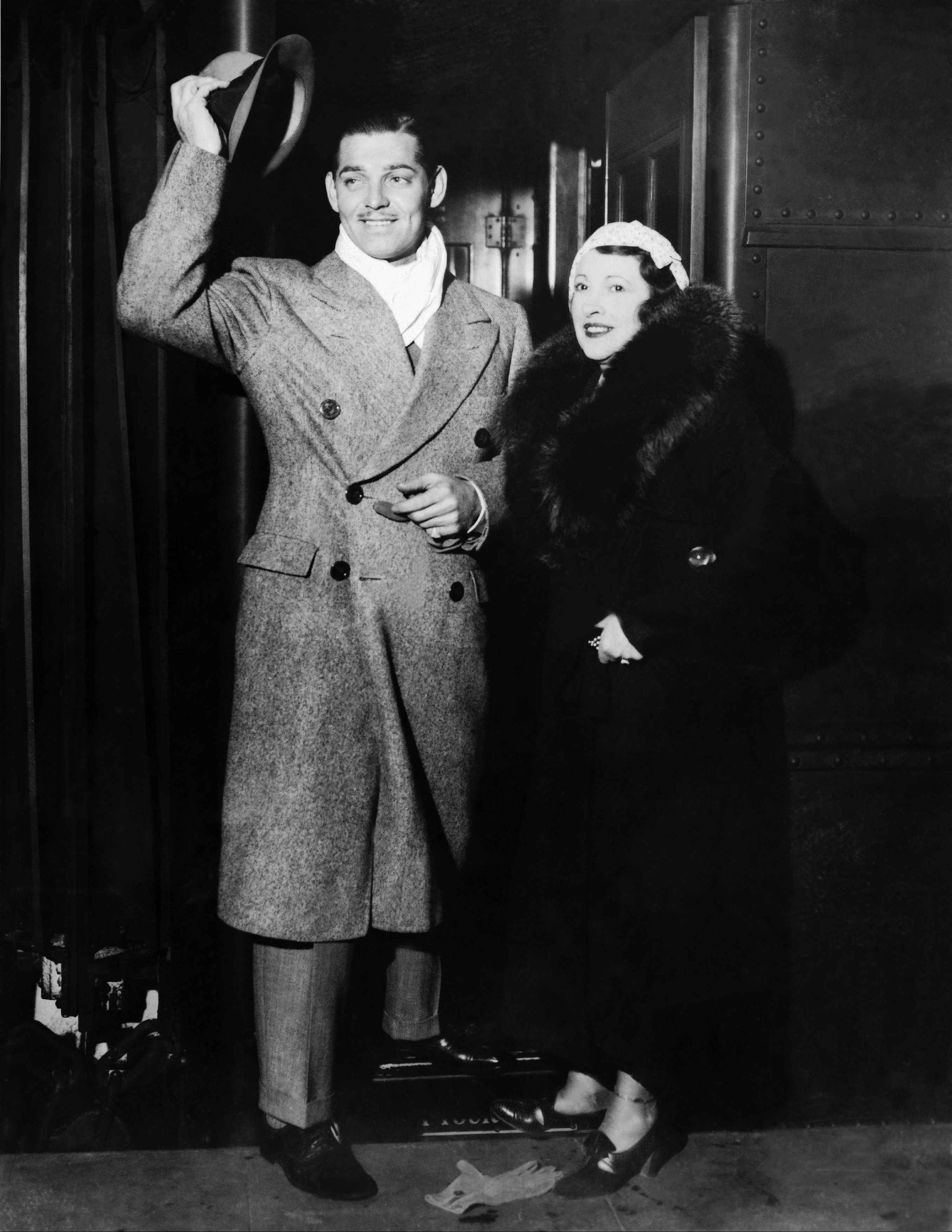 Clark Gable in a tailored coat and hat tips his hat, standing beside his wife Maria Langham in a fur-trimmed coat, both smiling and posing for a photo