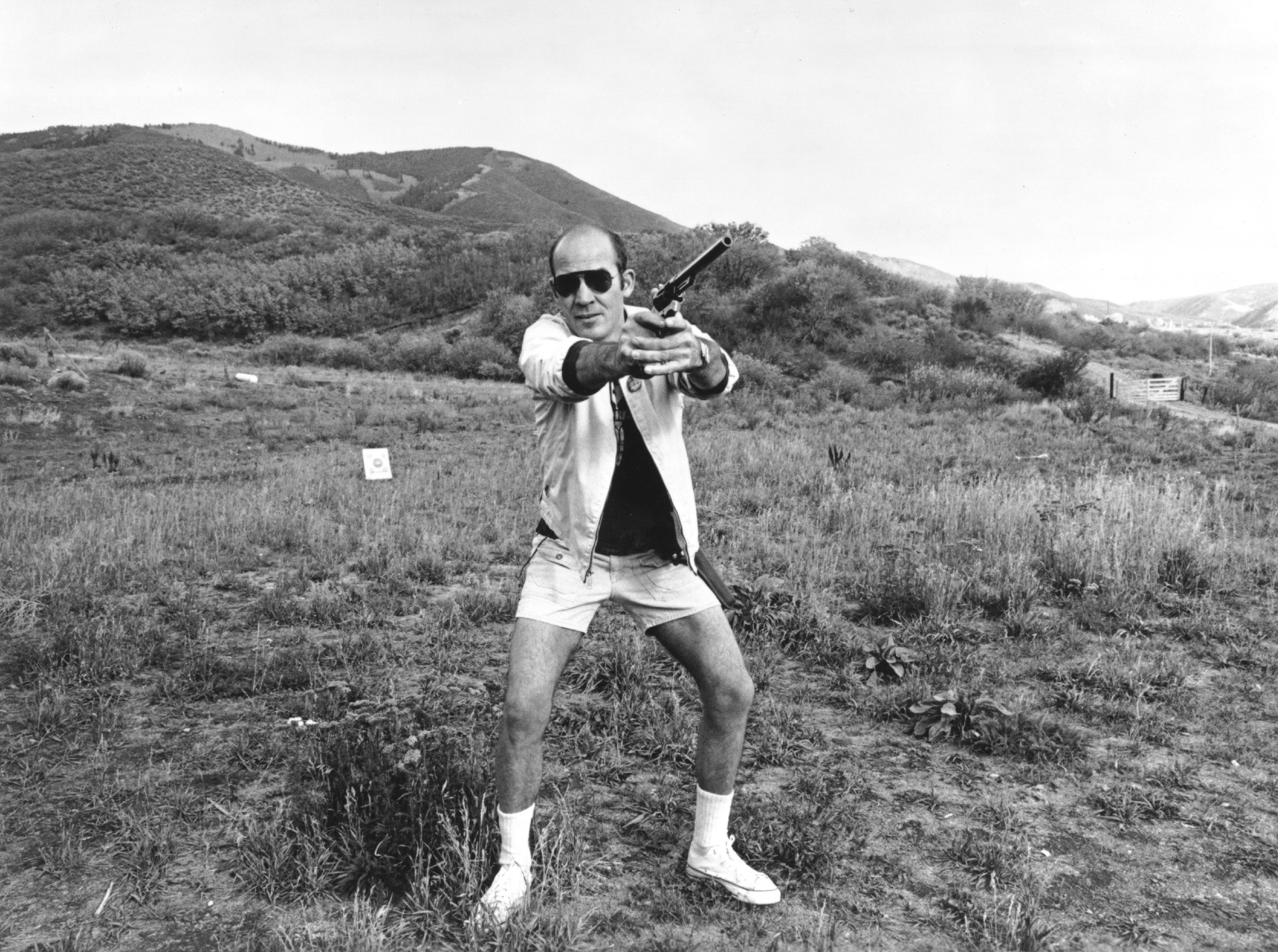 Hunter S. Thompson in sunglasses and casual wear poses outdoors with a pistol aimed forward, surrounded by grass and hills in the background