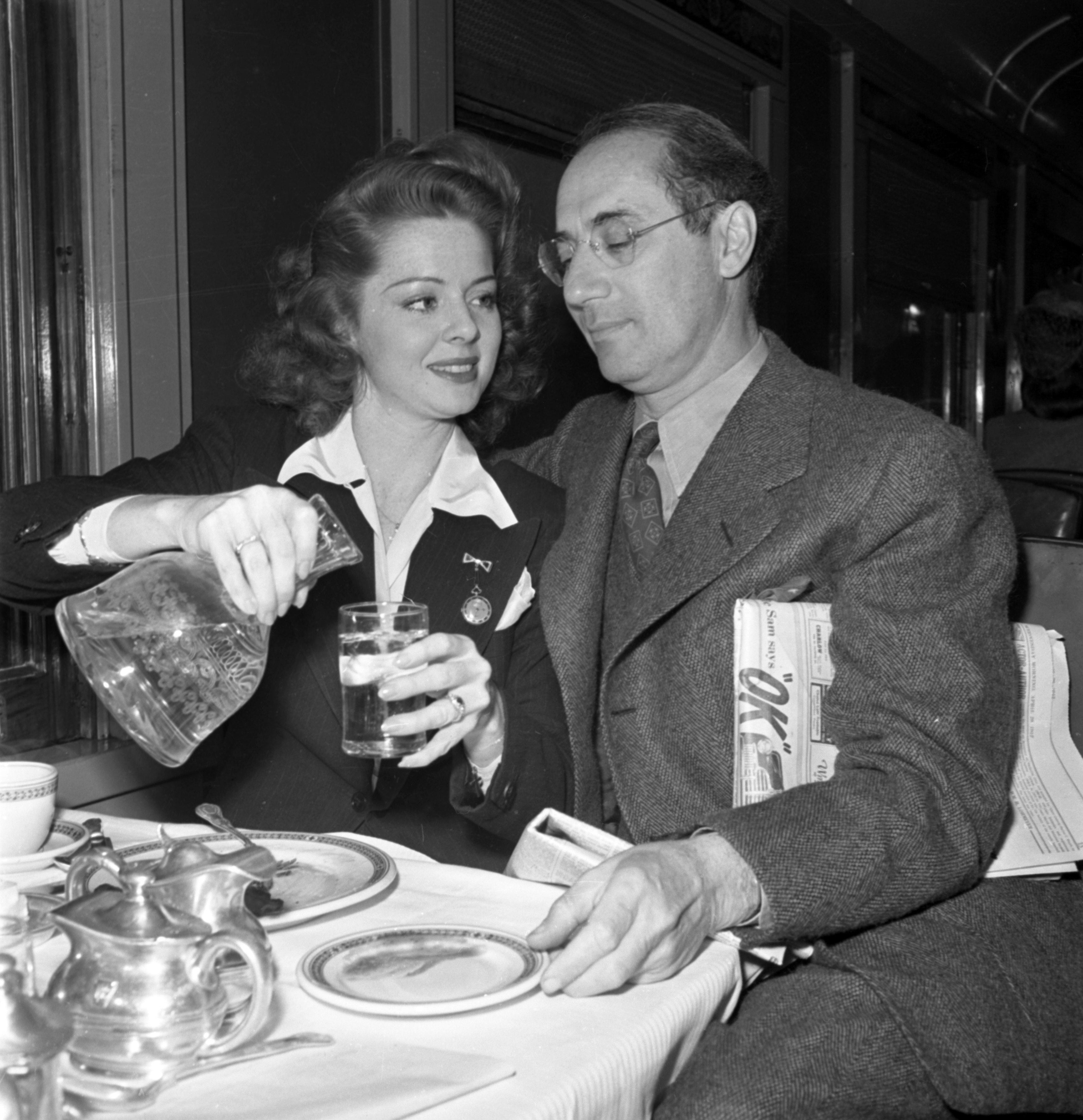 A woman elegantly pours water for a Groucho Marx at a table set with teacups and plates. The man holds a newspaper under his arm.
