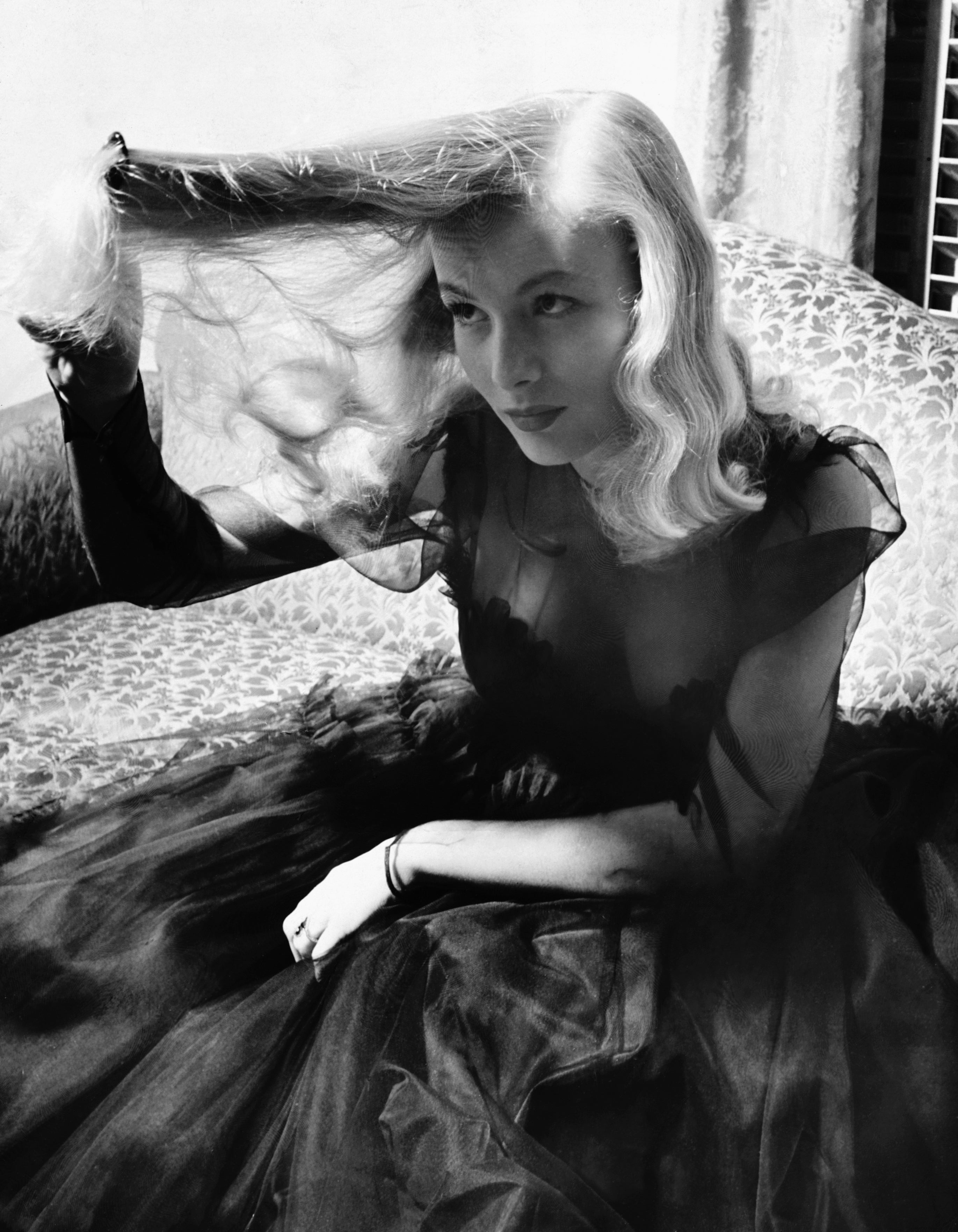 A blonde Veronica Lake in a vintage dress playing with her long hair, holding it up over her head, looking at it