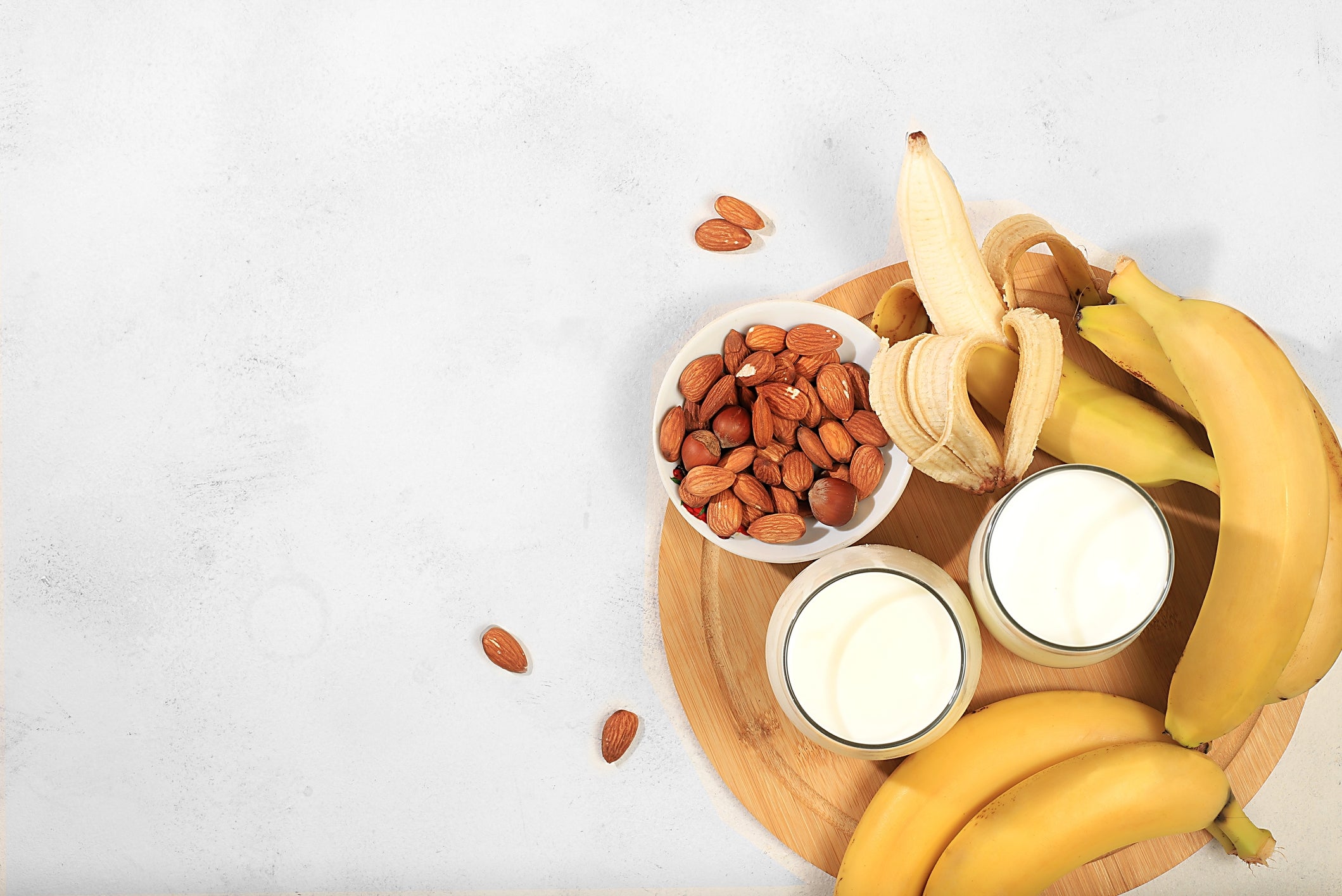 Bananas, peeled and whole, with almonds in a bowl, and two glasses of milk on a wooden tray