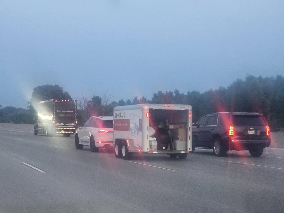 Vehicles driving on a highway; a U-Haul trailer is open with a person sitting inside amidst boxes