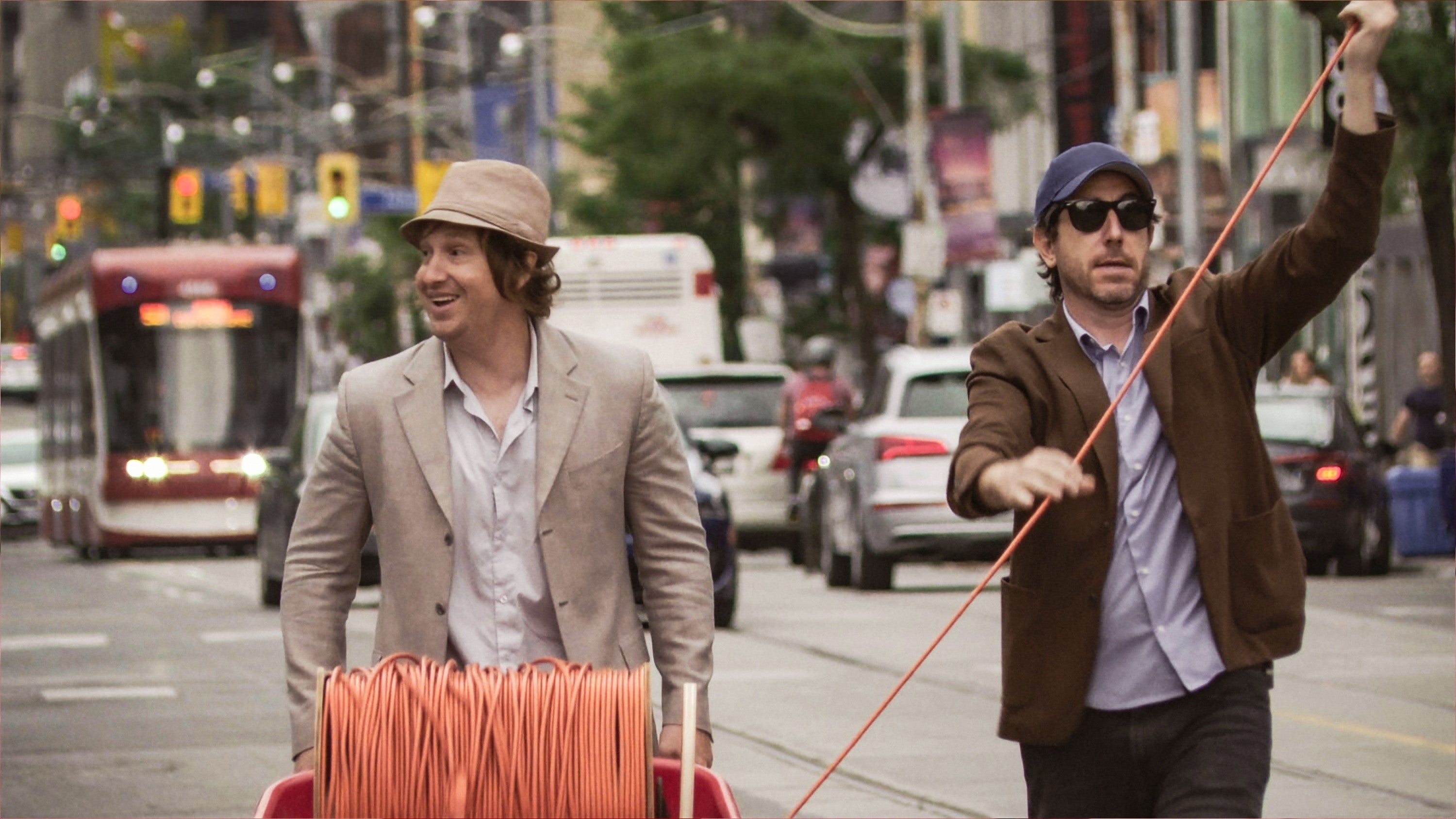 Two men on a city street pull an orange cable from a spool. One wears a beige hat and blazer, the other a blue cap and brown jacket
