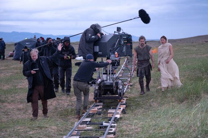 Film unit  operates camera connected  tracks outdoors. Two actors successful  play  costumes locomotion  alongside, surrounded by vast, grassy landscape