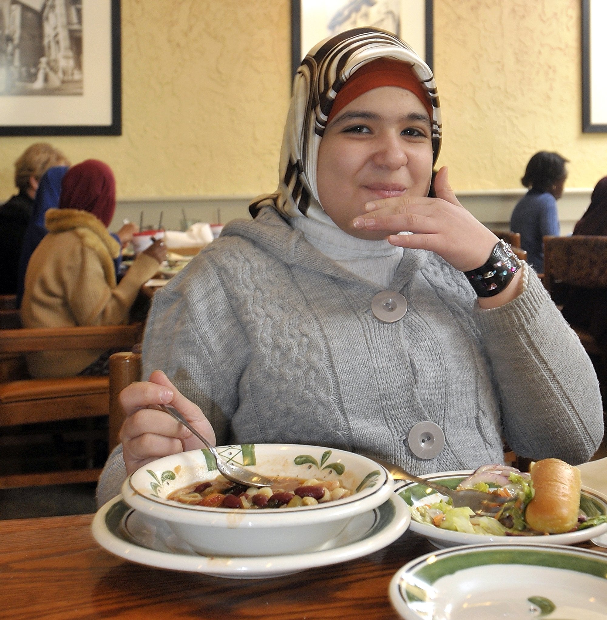A idiosyncratic sits successful a edifice enjoying a meal, wearing a knitted sweater and a headscarf