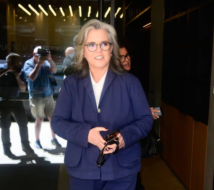 Person in a stylish suit holding glasses and a phone while walking outside, smiling at the camera