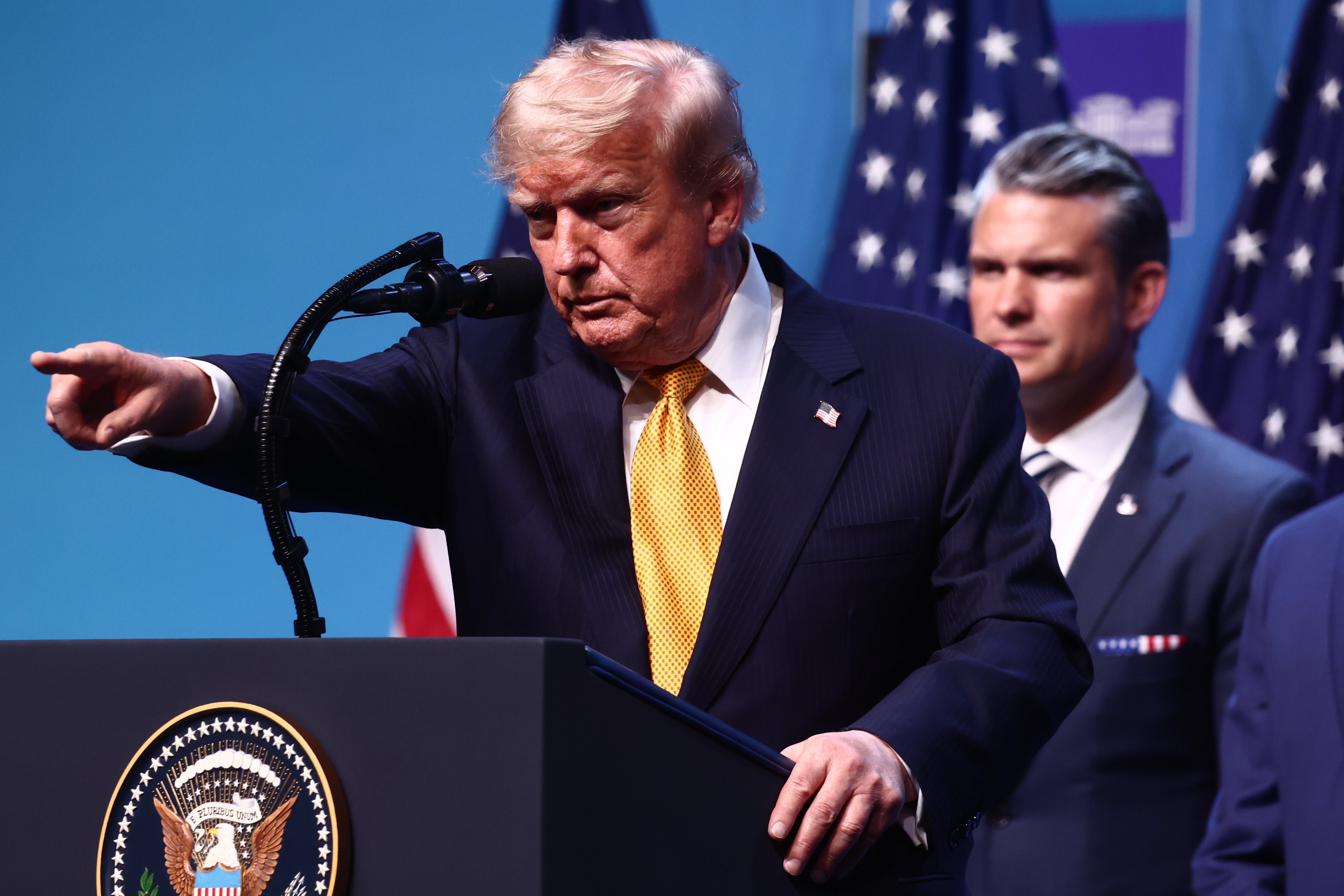 Man in a suit speaking at a podium with the presidential seal, pointing. Another man stands in the background. Flags are visible behind them