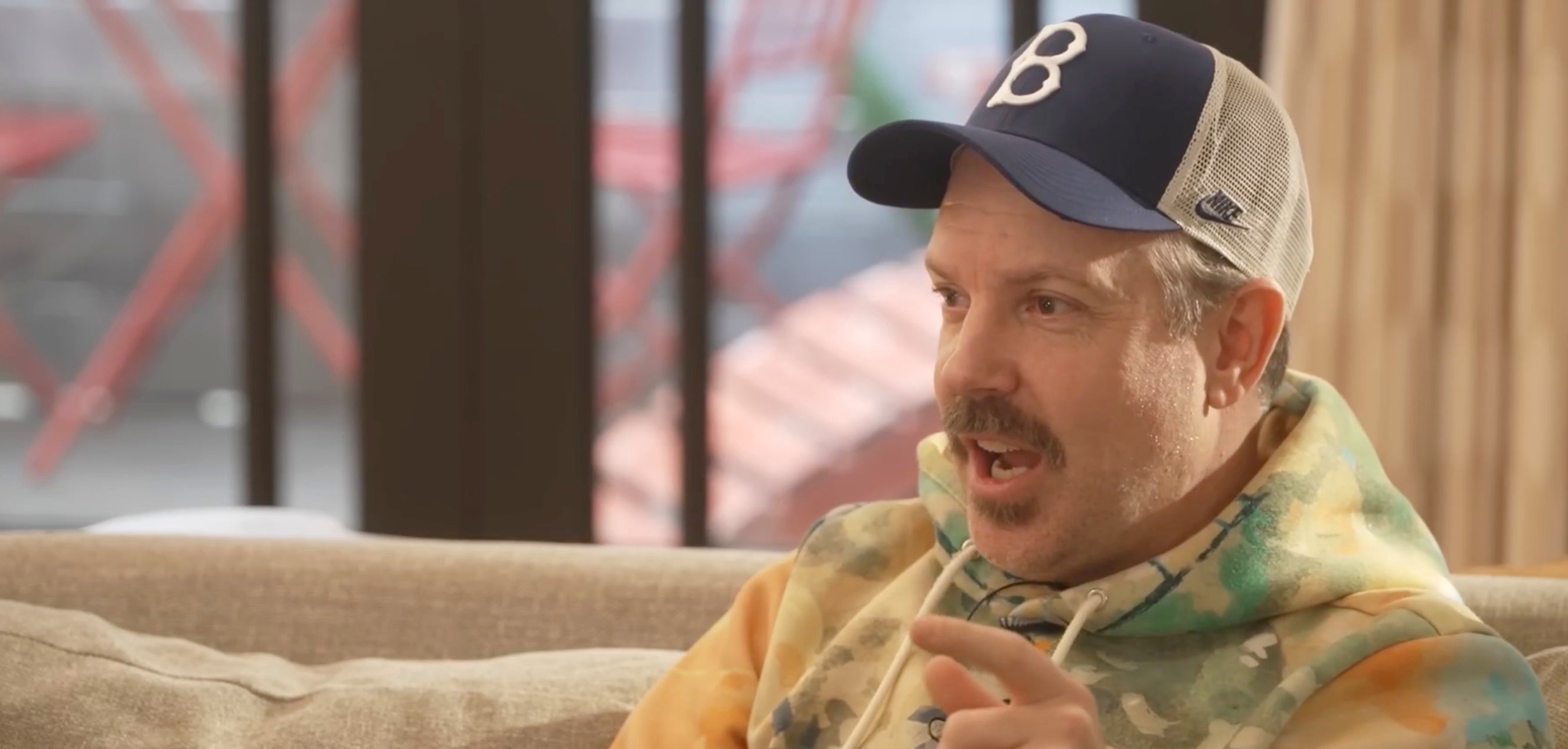 A person wearing a baseball cap and patterned hoodie sits on a couch, speaking animatedly