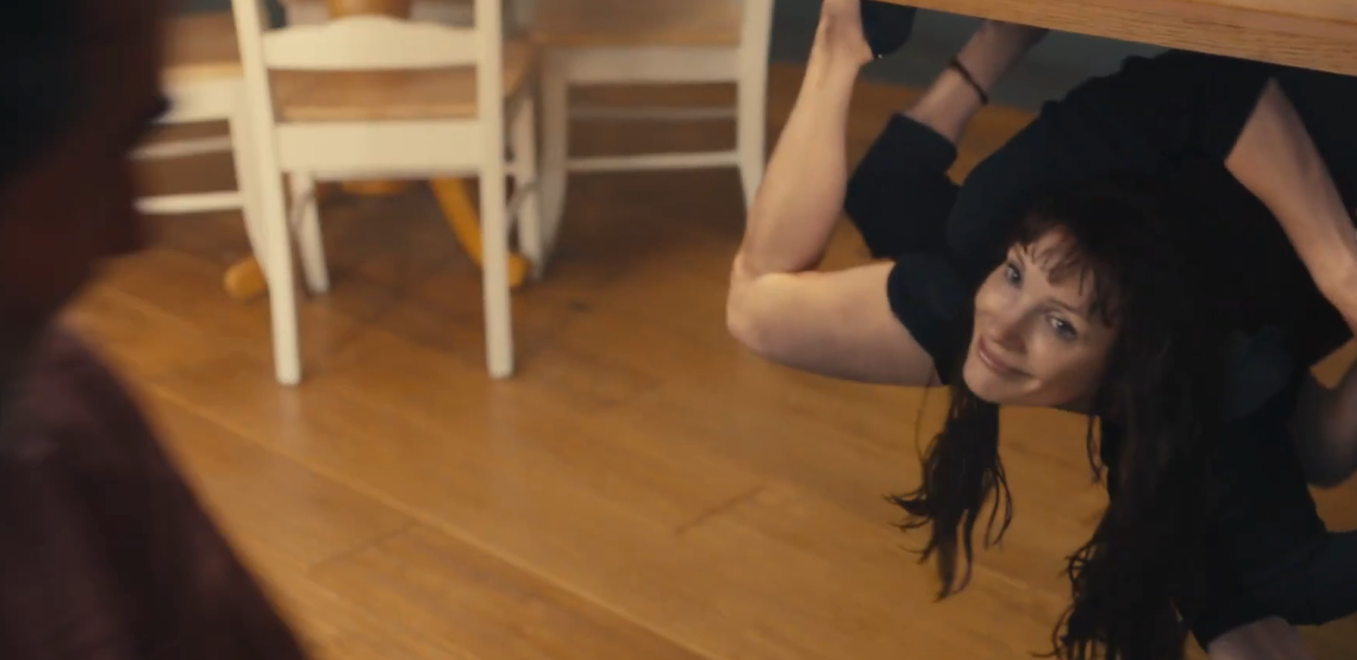 Person under a table playfully leans sideways, resting on their back, smiling at someone off-camera in a domestic setting