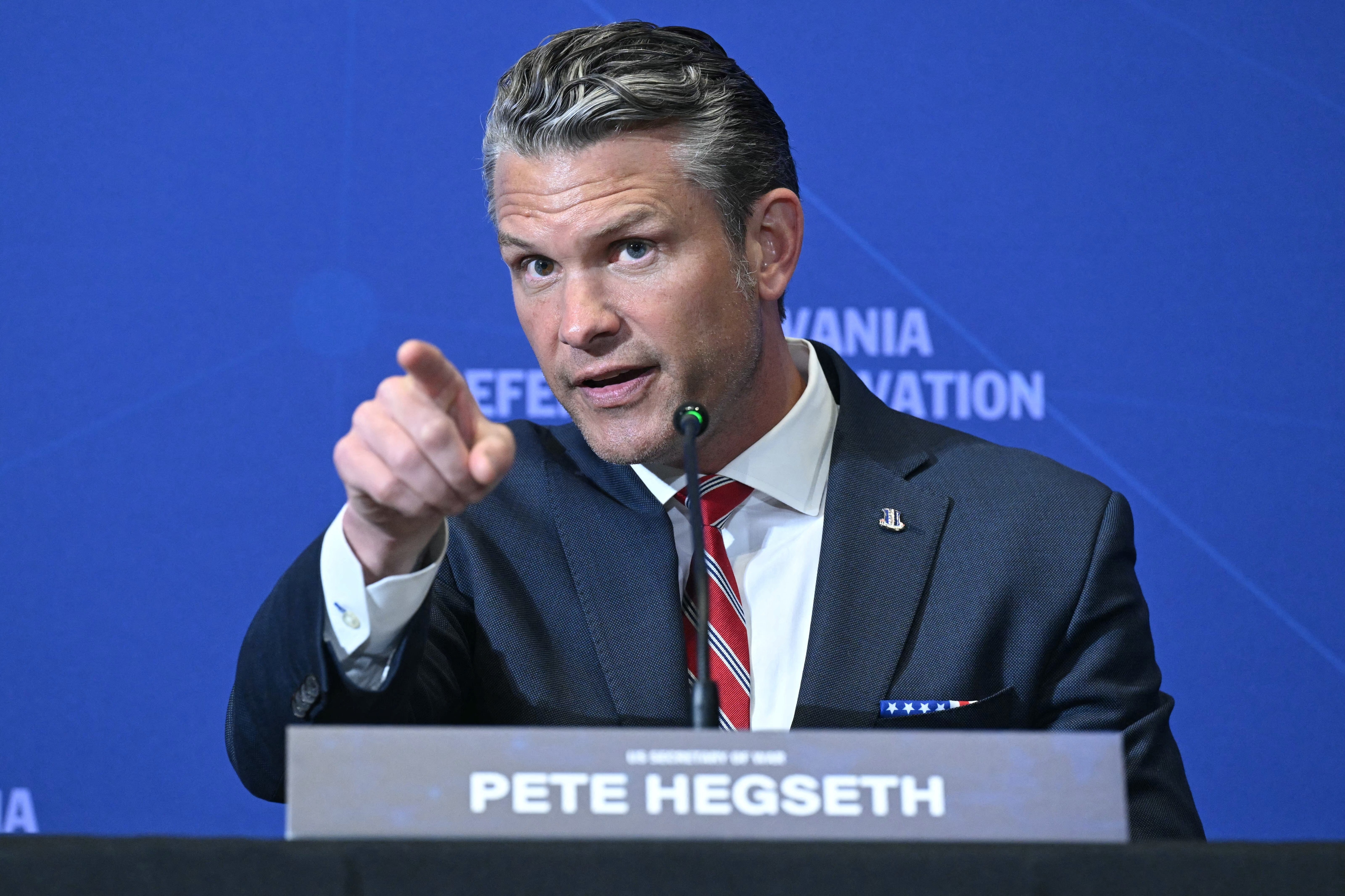 A person in a suit speaks at a conference, gesturing emphatically. A nameplate reads "Pete Hegseth."