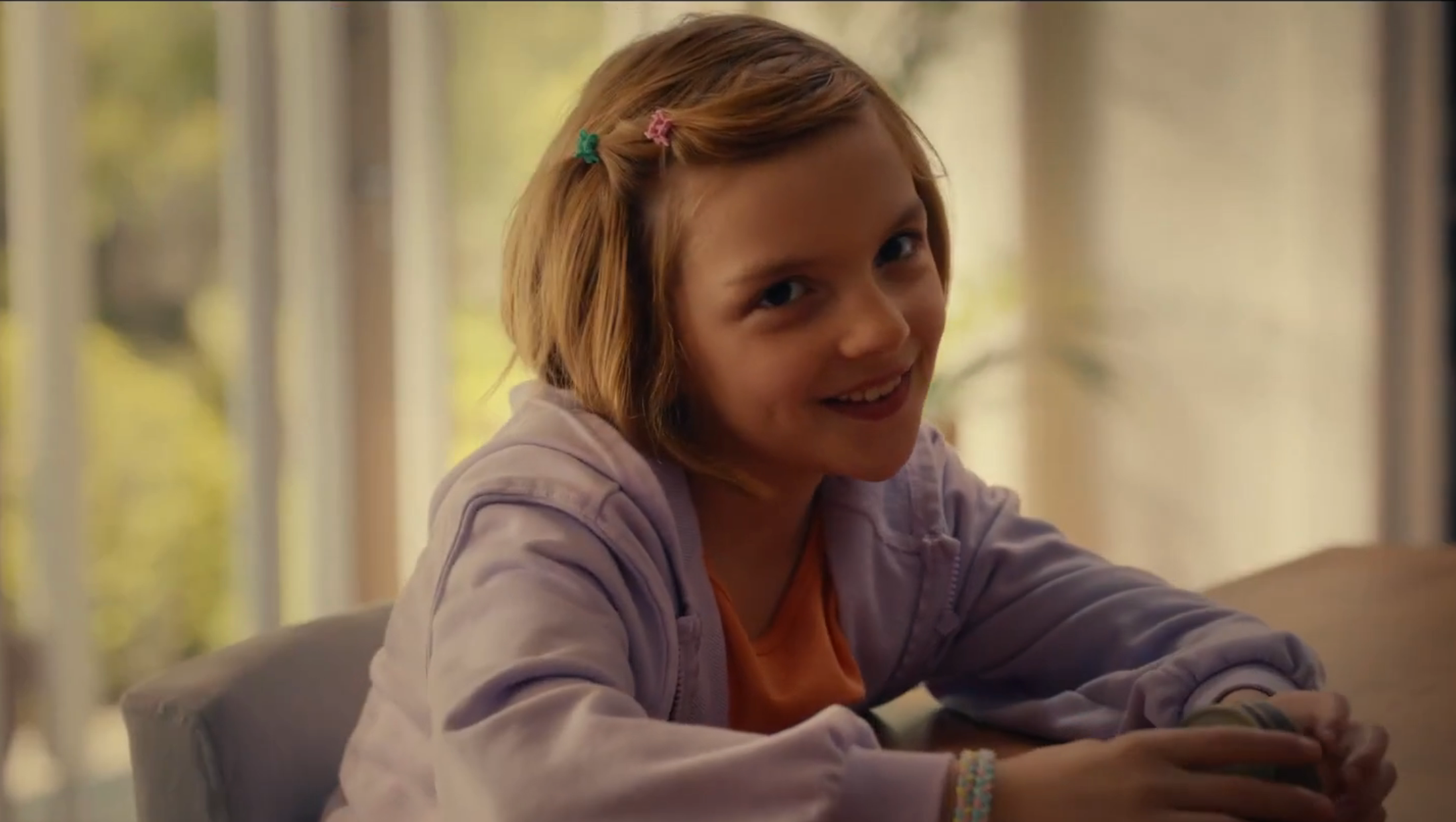 A young girl with short hair, wearing a casual hoodie, smiles at the camera indoors