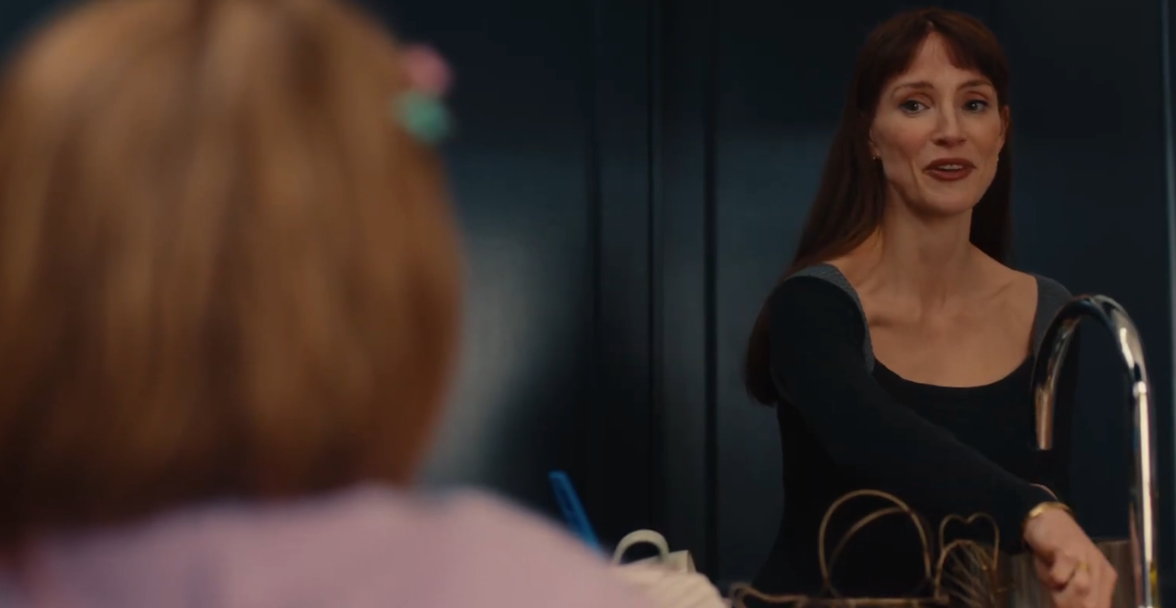A woman in a black outfit is smiling and gesturing while speaking to someone off-screen in a casual setting