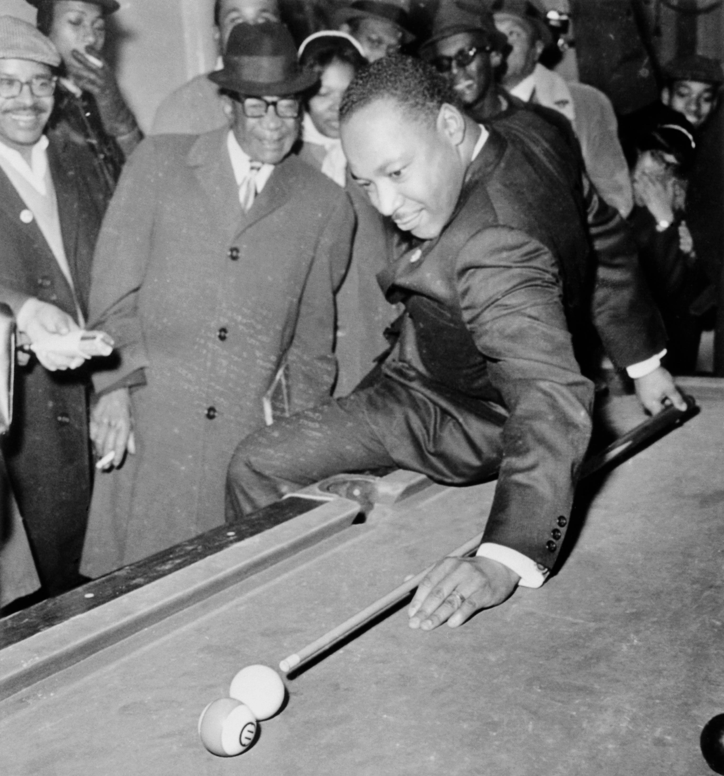 Martin Luther King in a suit playing pool, leaning backwards over the table, surrounded by onlookers in suits and hats, indoors
