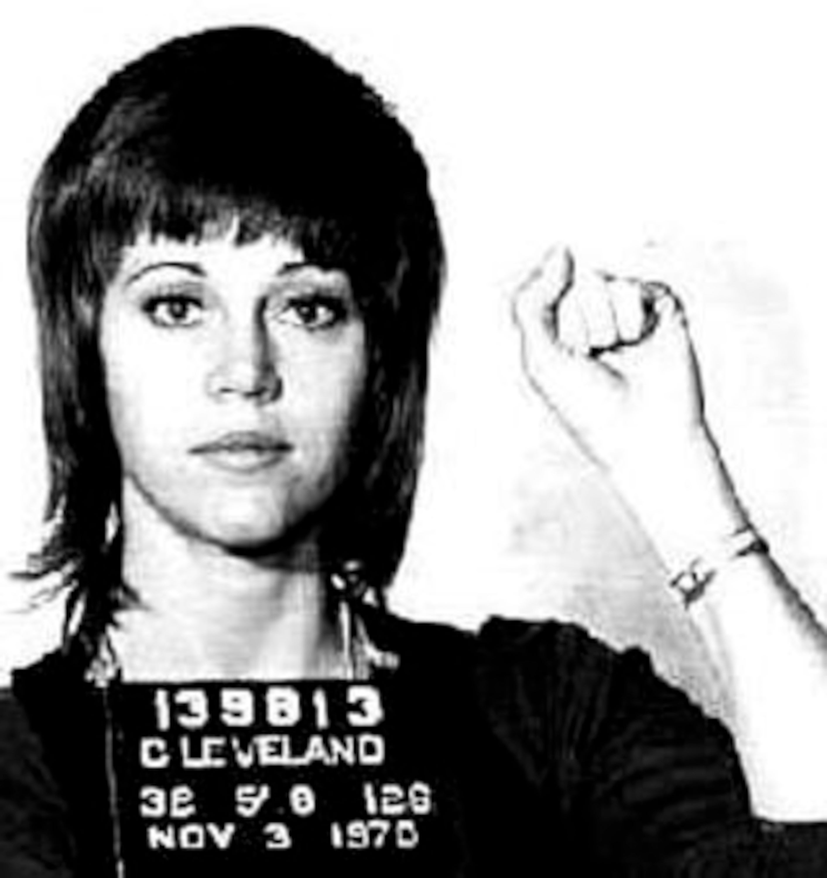 Jane Fonda with a short haircut holds up a fist in a police mugshot from 1970