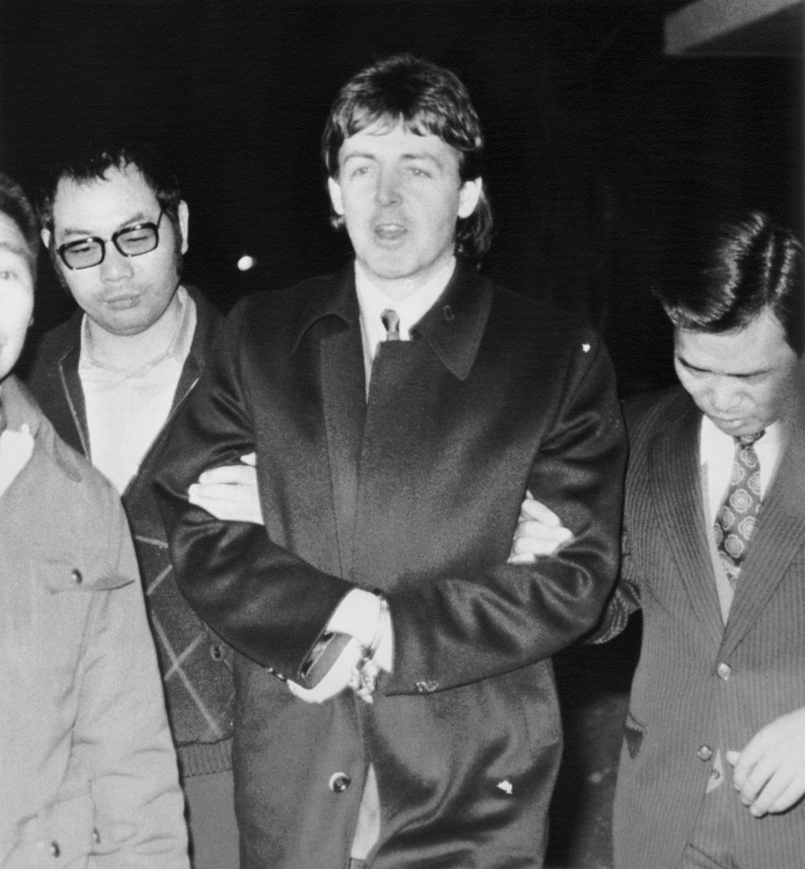 Paul McCartney in a coat is escorted by two others in suits, appearing to be in a serious or tense situation