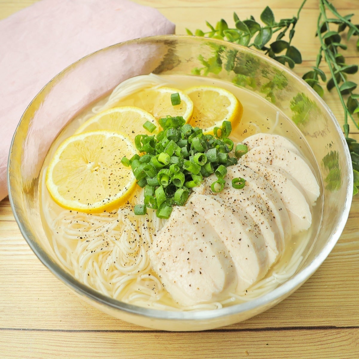 レモンとネギが乗った鶏肉のラーメンが透明なボウルに入っている。背景に植物が見える。