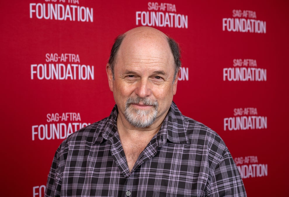 A person smiles in front of a SAG-AFTRA Foundation backdrop, wearing a plaid shirt
