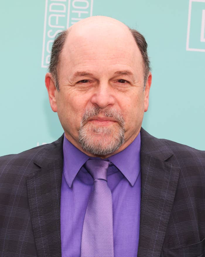 A man in a checkered suit with a purple tie stands on a turquoise backdrop at an event