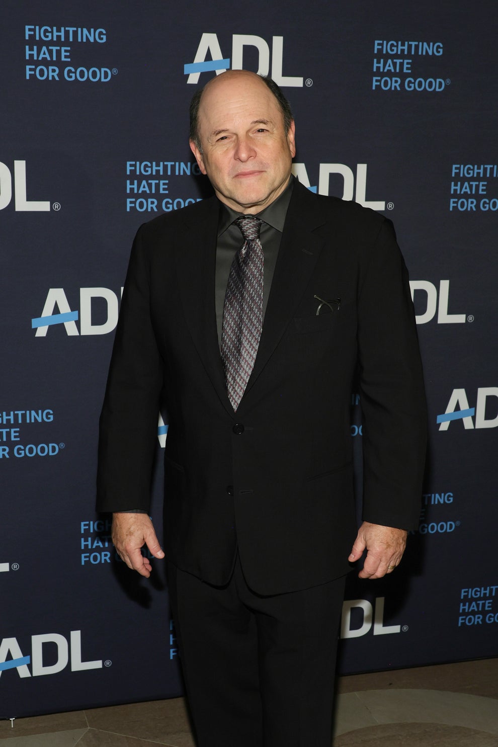 Person in a black suit and tie poses at an event with "Fighting Hate for Good" banners in the background