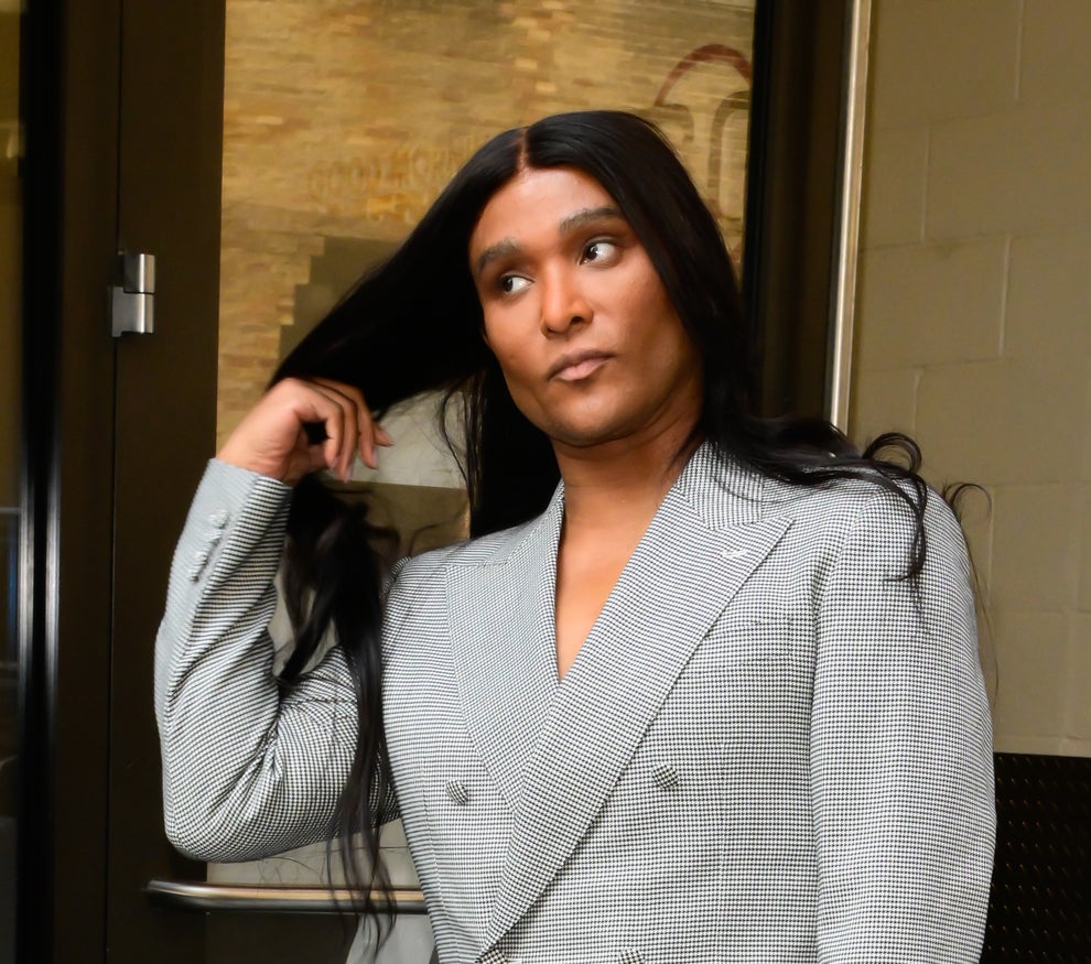 Person with long hair in a checkered suit, brushing their hair back with a confident expression