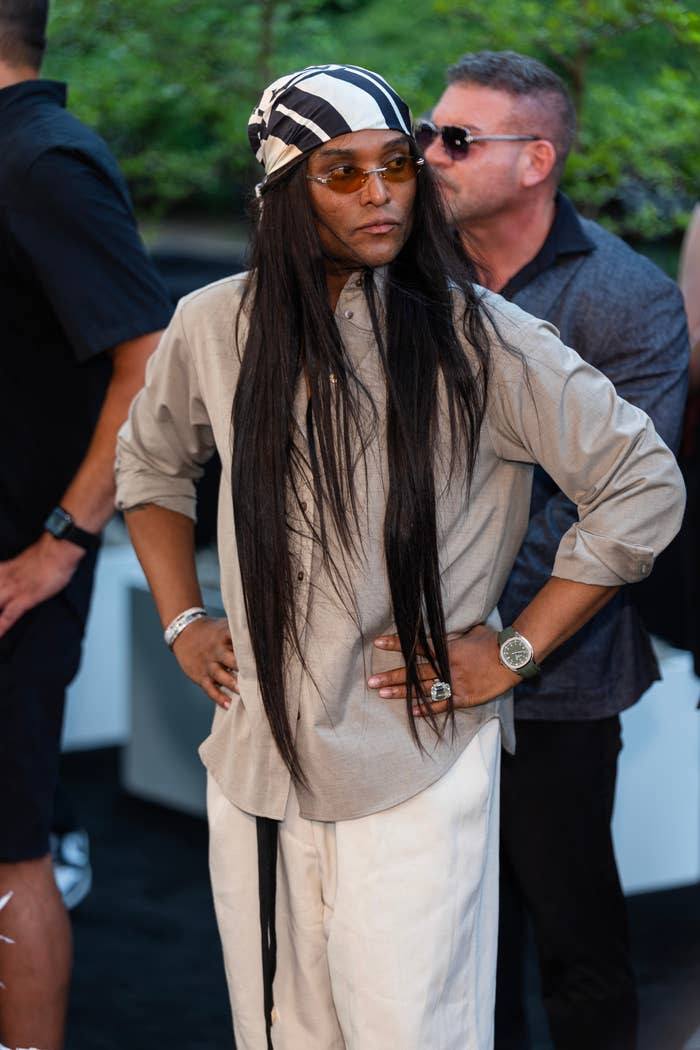 Person wearing a bandana, sunglasses, loose shirt, and pants at an outdoor event, standing confidently with hands on hips