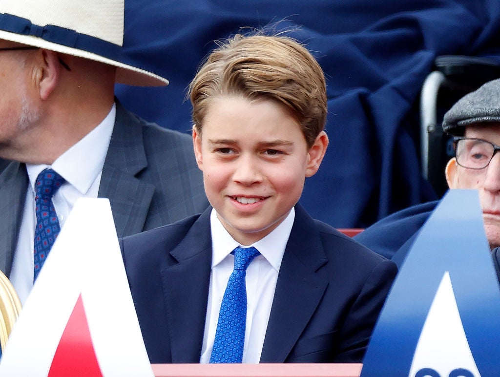 Young boy in a suit and tie smiles while seated at an event, surrounded by people, including someone in a suit and a hat