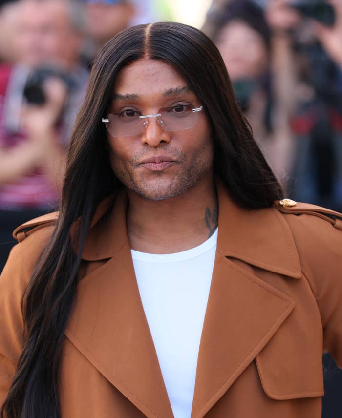 A person with long hair and glasses is wearing a stylish brown coat over a white shirt, walking in a public setting