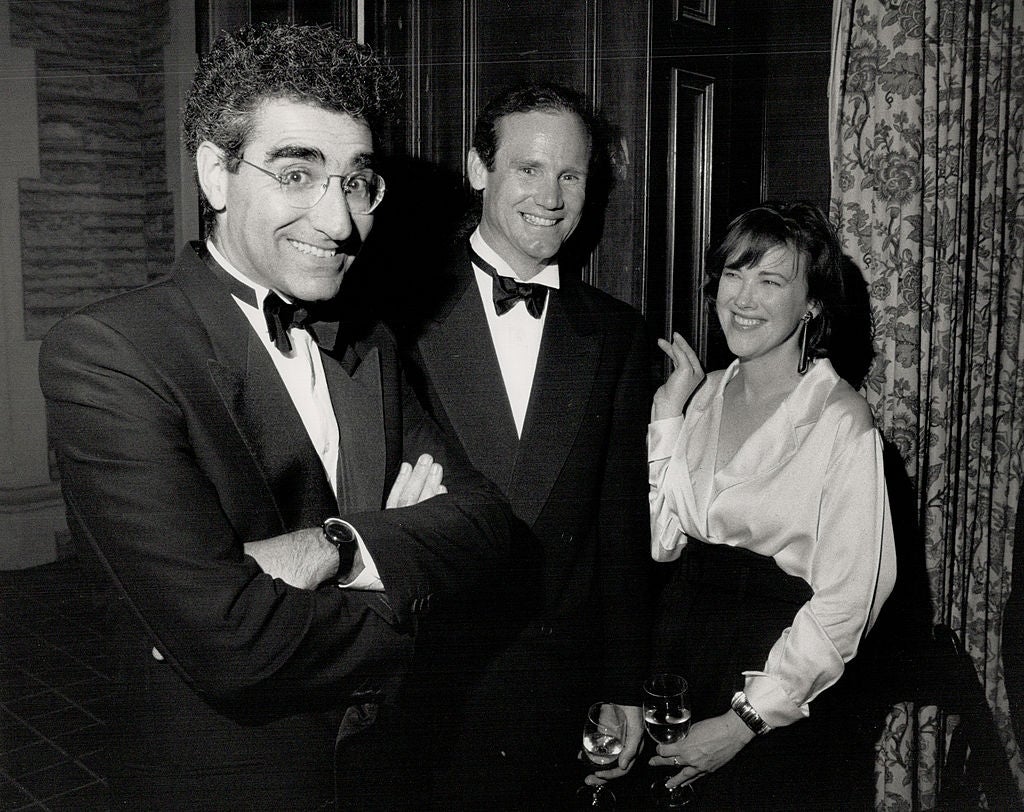 Three people in formal evening wear at an event. Two men are in tuxedos, and a woman is in a blouse and skirt, holding a drink, smiling