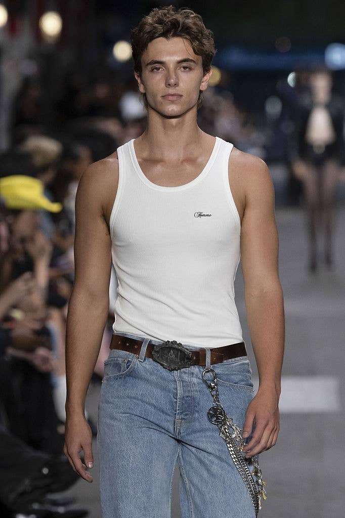 Person walking on a runway, wearing a white tank top and blue jeans with a chain belt