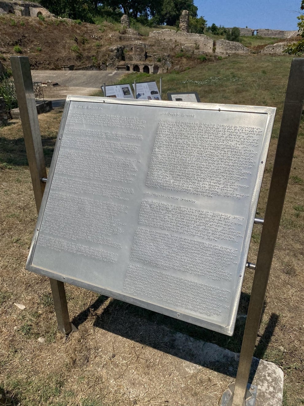 Metal display panel with Braille and print text about ancient ruins, situated outdoors on a grassy area with trees and ruins in the background