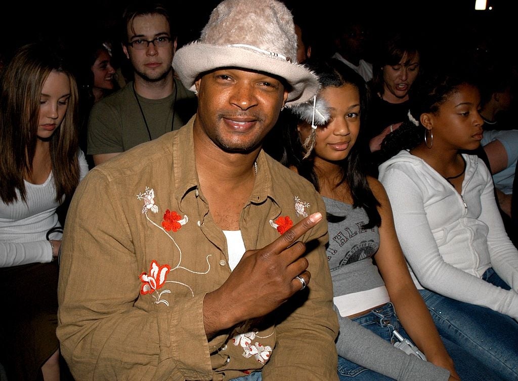 Person at an event wearing a floral embroidered shirt and light hat with a feather, sitting among a group of people