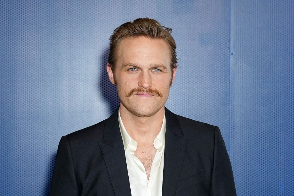 Man in a suit with a white shirt and mustache, standing against a plain backdrop