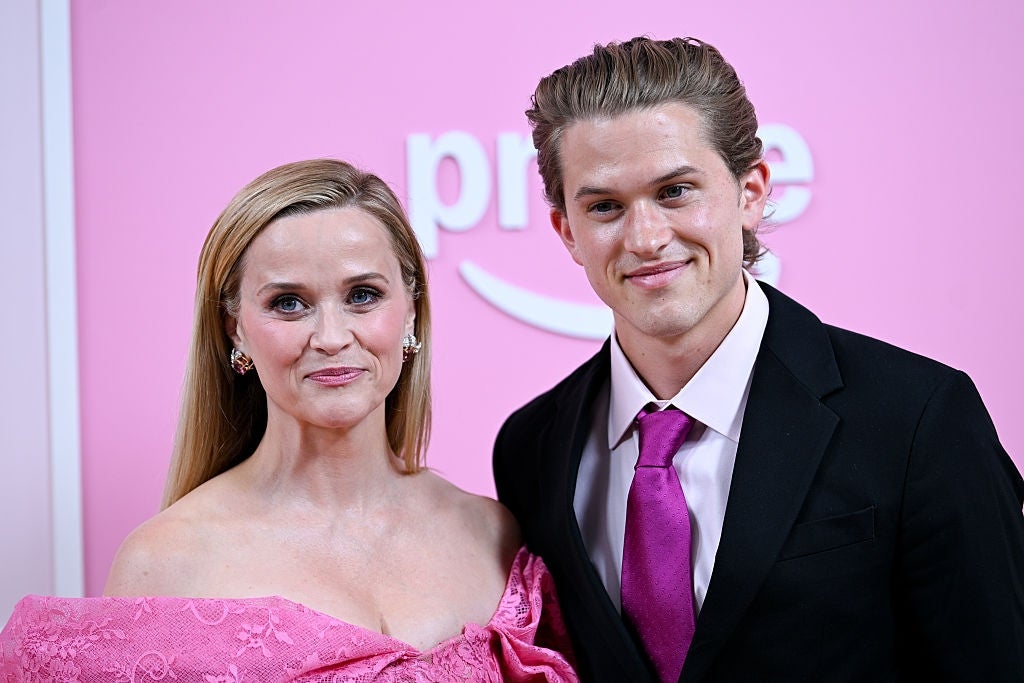Two people pose together on a red carpet. The woman wears an off-shoulder gown, and the man wears a suit with a tie. They are smiling